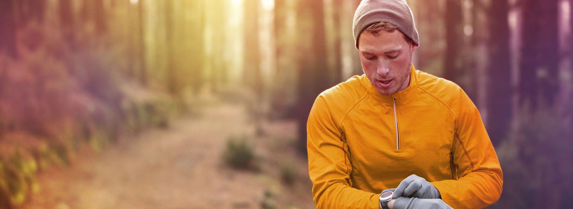 Ein Mann mit orangenem Pullover, Mütze und Handschuhen schaut beim Joggen im Wald auf seine Pulsuhr