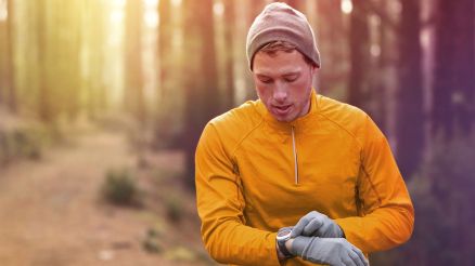 Ein Mann mit orangenem Pullover, Mütze und Handschuhen schaut beim Joggen im Wald auf seine Pulsuhr
